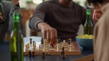 Cheerful man playing board games with group of friends, enjoying chess match competition at home. Young adult having fun with people, drinking beer and eating snacks. Handheld shot. Close up.