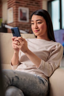 Asian woman smartphone user at home on couch, looking at phone with authentic smile. Internet web technology mobile user
