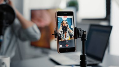 Social media famous influencer recording video blog with modern smartphone while speaking to audience. Joyful digital content creator sitting at studio desk while filimg vlog. Close up