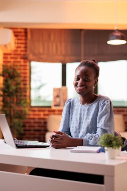 Female african american smiling student sitting at modern workplace desk in home office. Confident young professional freelancer working on computer and looking at camera