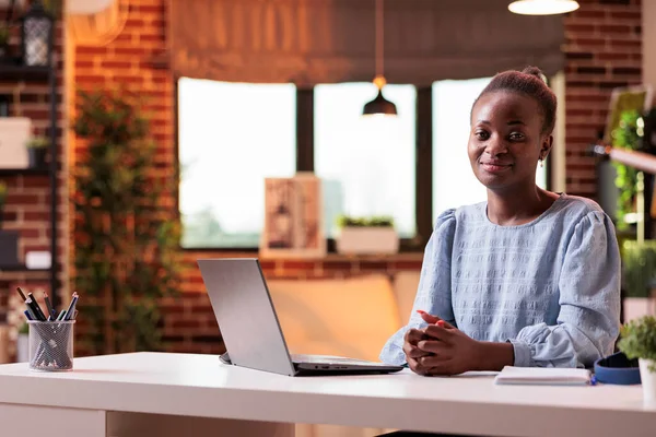 Female remote worker sitting near open laptop and looking at camera in modern home office. Young successful african american businesswoman at workplace in contemporary room