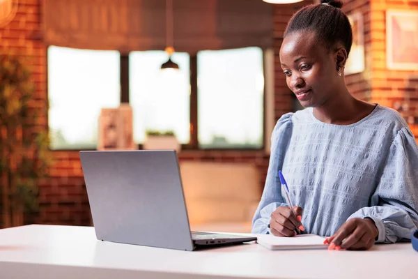 Woman studying with laptop and taking notes in notebook. Female african american remote university student watching educational tutorial and learning with online courses on internet