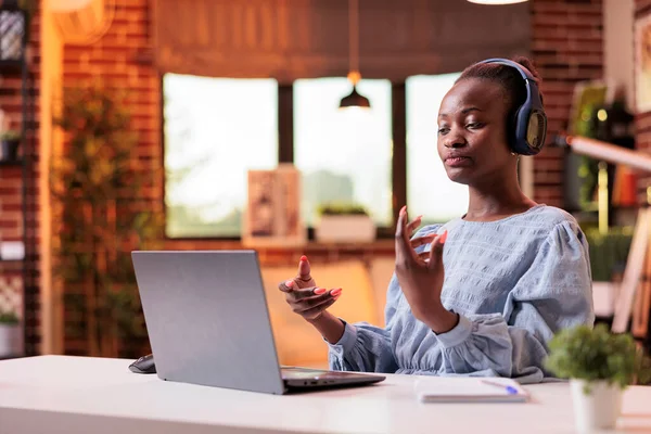 African american woman having video conference with coworkers while sitting in modern home office. Female remote worker wearing headphones and talking on online meeting in room with warm sunset light