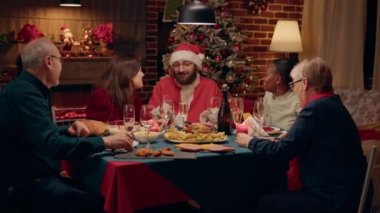 Joyful man disguised as Santa Claus clinking champagne glass with family enjoying Christmas dinner. Festive relatives celebrating winter feast while sitting at table with Santa Claus looking person.
