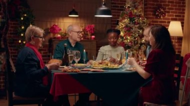 Happy senior man inviting family members to toast while celebrating winter holiday. Festive relatives gathered inside cozy apartment with Xmas authentic decorations to celebrate Christmas together.