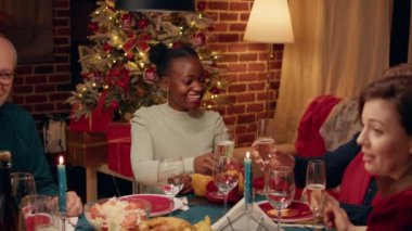 Woman at Christmas dinner table clinking glasses with family member while smiling at camera. People in festive living room celebrating winter traditional holiday inside ornate cozy apartment.