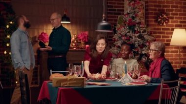 Festive diverse people at home enjoying Christmas dinner together. Joyful multiracial family members celebrating winter feast inside cozy apartment decorated with authentic Xmas ornations.