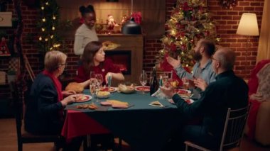 Beautiful woman bringing main course to Christmas dinner table. Cheerful festive family members gathered inside Xmas decorated apartment to celebrate winter holiday with traditional home cooked food.