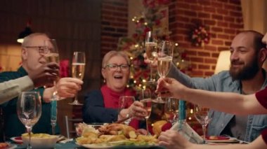 Happy family toasting sparkling wine at Christmas dinner table in living room. Multiethnic positive people gathered inside Xmas decorated apartment with cozy setting to celebrate winter holiday.
