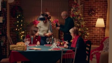 Positive senior talking with young man about rare bottle of wine brought at Christmas dinner table. Happy multiethnic women sitting in living room while celebrating winter traditional holiday together