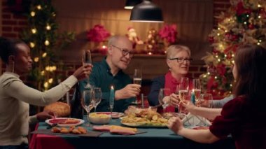 Festive diverse family members celebrating Christmas at evening dinner while clinking glasses with champagne. Cheerful close relatives enjoying winter holiday together. Handheld shot