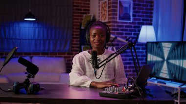 Evde kayıt stüdyosundaki masada oturan ve dizüstü bilgisayardan içeriği okuyan Afrikalı Amerikalı kadın portresi. Çevrimiçi radyo sunucusu canlı yayın yaparken kameraya kendinden emin bir şekilde gülümsüyor.