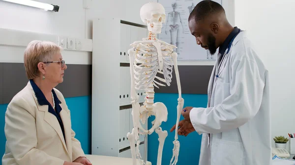 Health specialist examining human skeleton with senior patient in medical cabinet, doing anatomy bones examination, osteopathy spinal cord analysis. Osteopath at checkup visit with old woman.