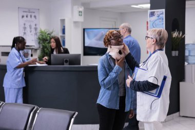 Specialist examining person with neck collar, wearing cervical foam brace after accident injury. Diverse people doing checkup visit consultation with medical appointment in waiting area lobby.