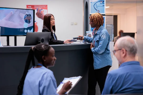 African american woman paying appointment with credit card transaction at hospital reception desk. Making electronic payment after doing checkup visit consultation at health center facility.