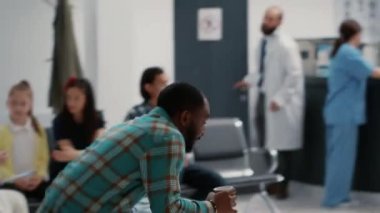 Busy hospital reception with diverse group of people and senior man with physical disability in waiting area. Patients and medical staff working on healthcare in clinic lobby. Handheld shot.
