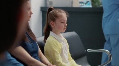 Portrait of little girl waiting with mother at hospital reception lobby, having medical consultation appointment. Small child sitting in waiting room area, healthcare examination.