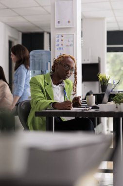 Employee in coworking open space, writing on clipboard, looking at laptop, sitting at workplace desk. Young african american woman working with company documents, taking notes