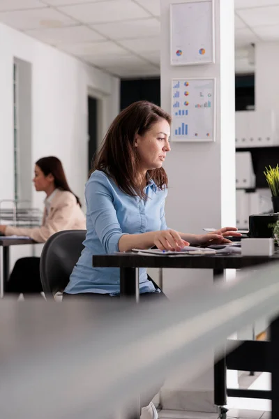 Serious young company employee working in coworking space, corporate worker sitting at workplace desk, side view. Attractive serious woman using laptop, manager in modern office