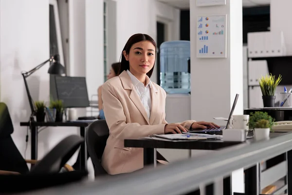 Employee working on laptop in coworking open space, company worker sitting at workplace desk portrait. Smiling attractive asian woman looking at camera in modern corporate office