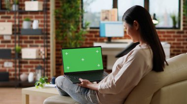 Young woman sitting at home with laptop having green screen chroma key display. Adult person sitting on sofa with portable computer on lap having mockup isolated template background. Tripod shot