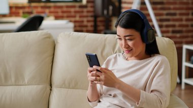 Smiling woman sending text messages on mobile phone while listening to music and doing work from home. Happy young adult person chatting with friend on smartphone while working remotely.