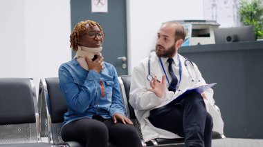 Female patient with cervical neck foam talking to doctor in waiting area at healthcare facility. Hurt woman in pain with collar brace trying to cure physical injury, help from medic. Handheld shot.