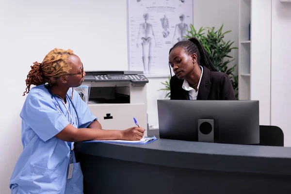 African american medical team analyzing report forms with patient insurance support on papers. Nurse and receptionist working on checkup visit appointments at hospital reception desk, healthcare help.