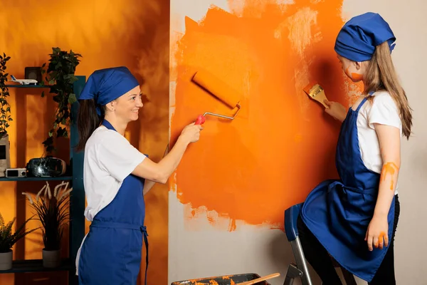 Small family using orange paint on walls, young child on ladder painting surface with paintbrush and mother using roller brush tool. People having fun with housework renovation, home decor.