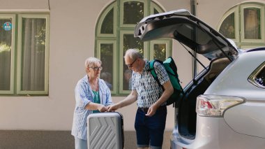 Retired couple loading voyage luggage in car trunk while going on marriage anniversary summer citybreak. Elderly tourists getting ready for field trip while putting baggage and trolleys inside vehicle