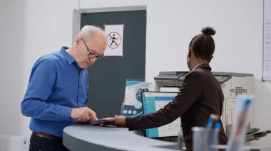 Old man using credit card to pay after consultation appointment, making payment transaction at hospital reception counter. Senior patent paying for checkup visit and treatment.