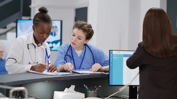 Diverse medical team working at hospital reception desk ith receptionist, analyzing checkup papers to help with healthcare appointments. Specialists writing insurance report at registration counter.