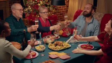 Multiethnic positive people gathered at home to celebrate winter holiday with home cooked traditional food and sparkling wine. Happy family toasting sparkling wine at Christmas dinner table.