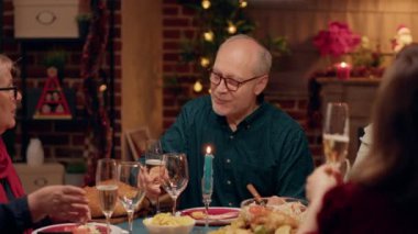 Happy senior couple talking to each other while enjoying Christmas dinner with loved ones. Elderly people sitting inside cozy warm apartment decorated with Xmas garlands and traditional dinnerware.