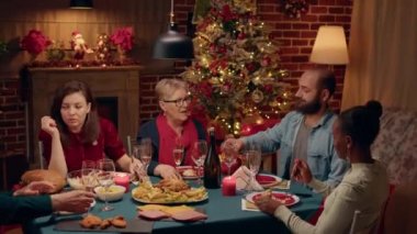 Confident man talking with mother-in-law at Christmas dinner while drinking champagne. Festive family gathered to celebrate winter holiday inside cozy apartment with fireplace and Xmas decorations.