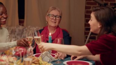 Happy senior woman sitting at table with relatives while enjoying Christmas dinner. Joyful elder person celebrating winter feast with close family members inside warm apartment with Xmas decorations.