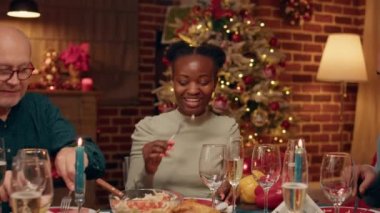 Multiethnic family members sitting at Christmas dinner table while enjoying seasonal home cooked food. Joyful relatives gathered inside cozy apartment decorated with Xmas ornations.