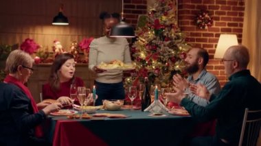 Festive african american woman bringing main course to table while people enjoying Christmas dinner. Joyful family members celebrating winter feast with traditional home cooked food.