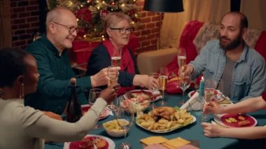 Festive family gathered at home to celebrate Christmas while toasting sparkling wine in dining room. Positive diverse people celebrating winter holiday inside cozy apartment with Xmas decorations.