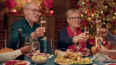 Cheerful close relatives enjoying winter holiday together while drinking sparkling wine. Festive people celebrating Christmas inside cozy apartment with traditional Xmas decorations. Tripod shot