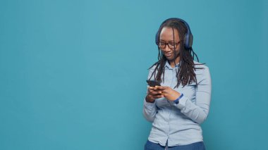 Female model using smartphone to listen to music on headset, having fun with radio song and dancing on camera. Looking at mobile phone and enjoying audio sound on earphones in studio.