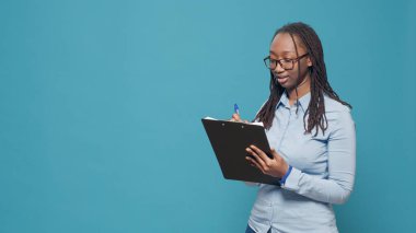 Portrait of young adult taking notes on textbook files, writing information on clipboard papers. Happy excited person feeling confident about text on paperwork documents in studio.