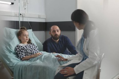 Pediatric therapist discussing with father of hospitalized sick daughter resting in patient bed. Doctor specialist examining ill kid under treatment resting in children healthcare facility ward.