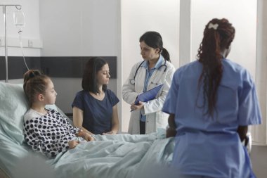 Pediatrician talking with mother about sick daughter health condition and disease evolution. Doctor specialist presenting treatment scheme to parent while ill little girl resting in patient bed.
