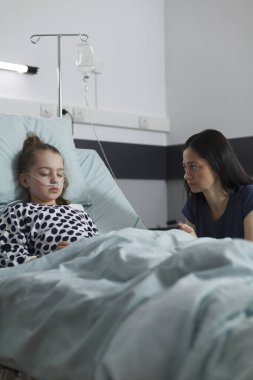 Ill kid under medicine treatment sleeping inside healthcare clinic pediatrics ward while parent waiting for her to wake up. Worried mother sitting beside sick daughter resting in patient bed.