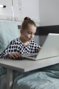 Ill kid resting on patient bed while enjoying gaming on modern computer. Sick little girl playing games on laptop while hospitalized in healthcare clinic pediatric ward room