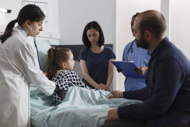 General practitioner consulting ill little girl under treatment inside clinic pediatric ward. Expert pediatrist checking sick girl health condition using stethoscope while parents sitting besider her.