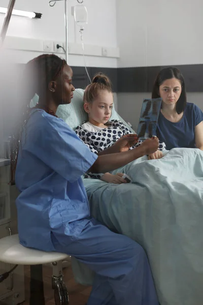 Pediatric healthcare facility staff analyzing MRI results of ill kid under treatment resting in patient bed. Nurse showing radiography scan image to sick girl while mother sitting beside her.