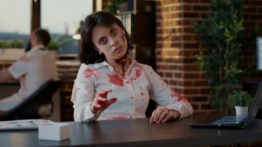 Weird looking spooky zombie woman waving at camera while sitting in office. Mindless brain-eating undead monster with bloody wounds sitting at table in workspace while waving hand.