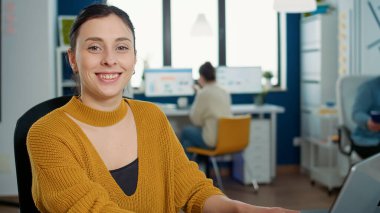 Portrait of woman typing using laptop keyboard and smiling at camera sitting at desk in busy startup office. Casual business employee working relaxed with papers with charts and statstics.